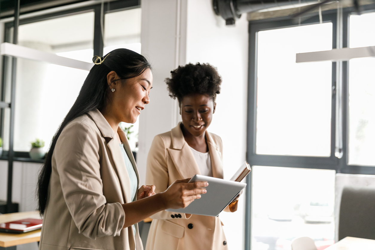 Two young businesswomen walking to a meeting together and looking at data on a digital tablet one of them is holding.